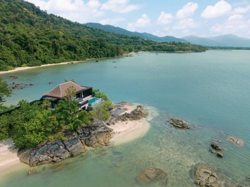 Tropisches Ufer mit Felsen, einer Strandhütte und grünen Bergen im Hintergrund.