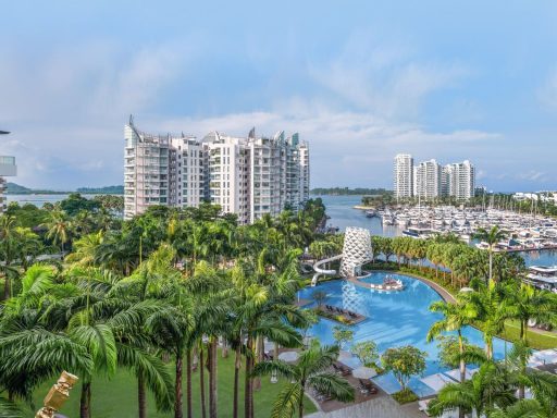Panorama mit modernen Wohngebäuden, Pool und Yachthafen am Wasser, umgeben von Palmen.