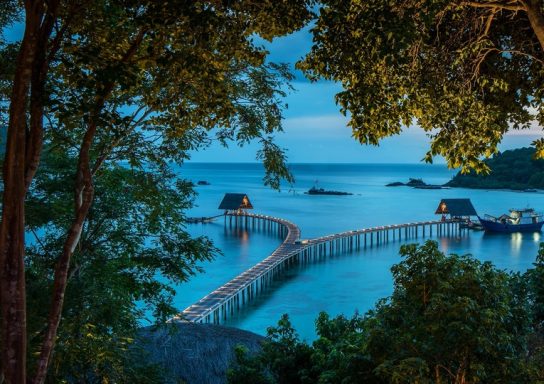 Ein Holzsteg führt über ruhiges Wasser zu zwei Hütten vor einer malerischen Küstenlandschaft.