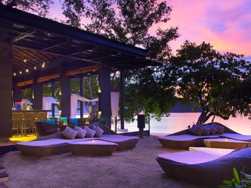 Tropischer Strandbereich mit Liegen, Bar und buntem Sonnenuntergang am Wasser.