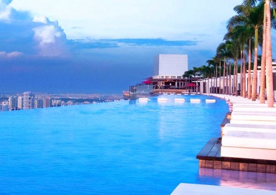 Infinity-Pool mit Blick auf die Stadt, umgeben von Palmen und einem Abendhimmel.