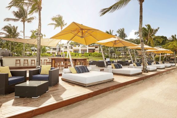 Helle Liegen mit Sonnenschirmen am Strand, umgeben von Palmen und einem Restaurant.