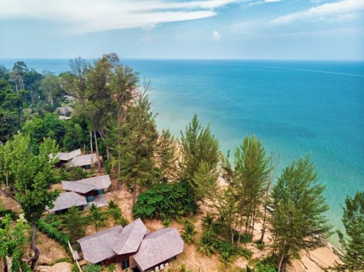 Küstenansicht mit Hütten, Strand und Palmen unter klarem Himmel.