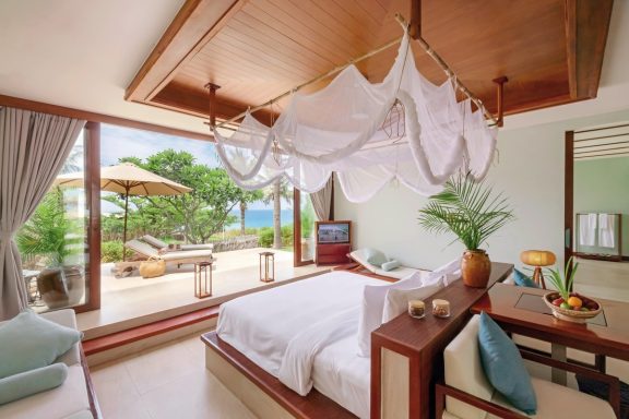 Luxuriöses Zimmer mit großem Bett, Holzmöbeln und Blick auf einen Garten und Strand.