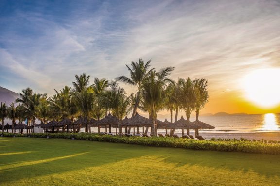 Tropischer Strand bei Sonnenuntergang, umgeben von Palmen und Liegen.