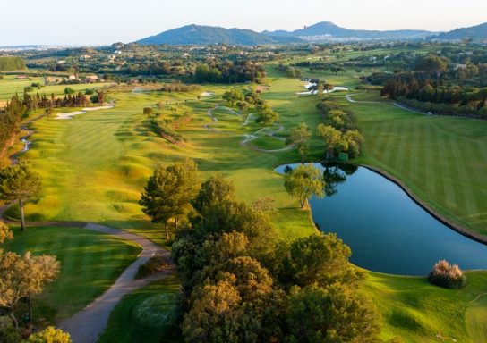 Landschaft mit Golfplatz, Bäumen, Wasser und Hügeln im Hintergrund.