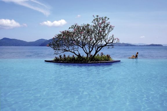 Ein ruhiger Pool mit einer kleinen Insel und Baum, umgeben von blauem Wasser und Bergen.