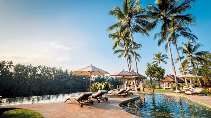 Tropischer Poolbereich mit Liegen, Sonnenschirmen und Palmen unter blauem Himmel.