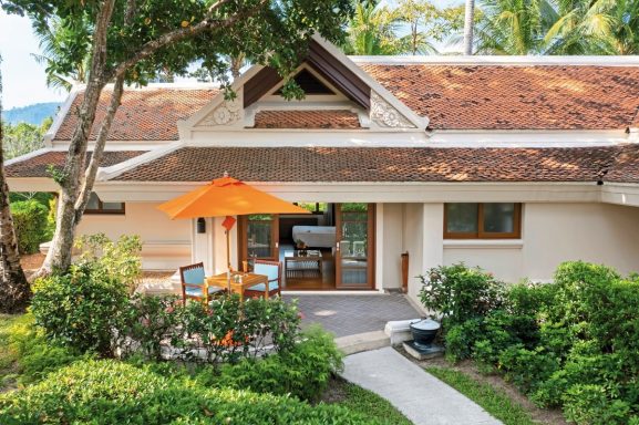 Ein helles Haus mit orangefarbenem Sonnenschirm, umgeben von üppigem Grün.