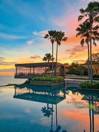 Sonnenuntergang über einem Pool, umgeben von Palmen und einem Strandhaus.