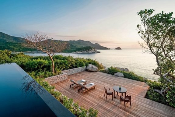 Blick auf eine Terrasse am Meer mit Sitzgelegenheiten und sanften Hügeln im Hintergrund.