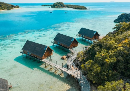 Drei Holzbungalows über klarem Wasser, umgeben von tropischer Vegetation.