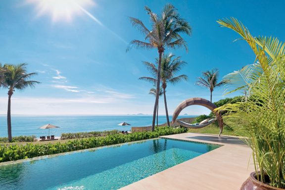 Luxuriöser Pool am Meer mit Palmen und Sonnenschein im Hintergrund.