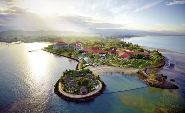 Tropische Insel mit Resort, umgeben von Wasser und grüner Vegetation, bei Sonnenaufgang.