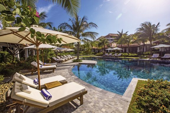 Poolbereich mit Liegen, Sonnenschirmen und Palmen umgeben von tropischer Vegetation.