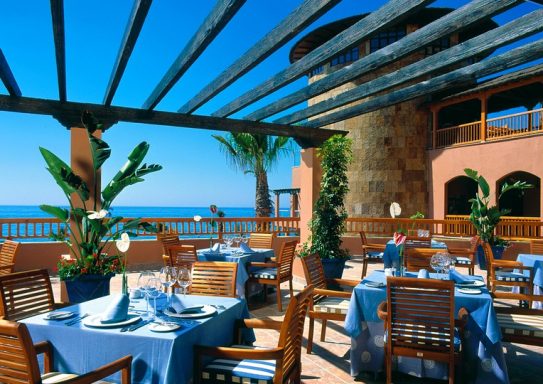 Gemütliches Restaurant mit blauen Tischdecken, Blick auf das Meer und Palmen im Freien.