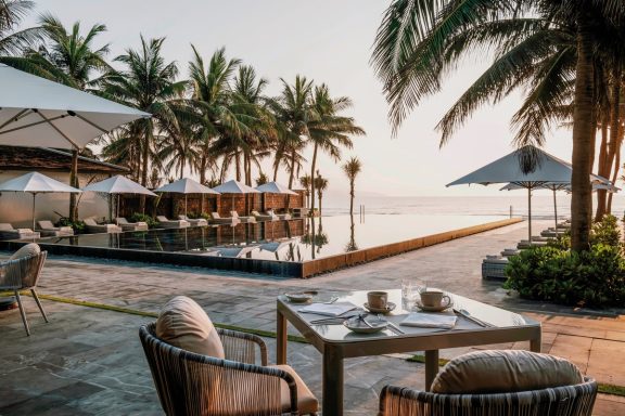 Luxuriöser Außenbereich mit Palmen, Pool und Essbereich am Wasser bei Sonnenaufgang.