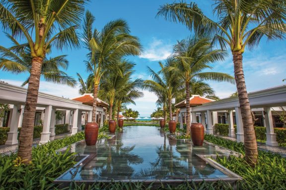 Tropischer Garten mit Palmen und reflektierender Wasserfläche zwischen Gebäuden.