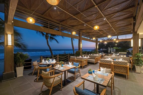 Gemütliches Restaurant am Strand mit gedeckten Tischen und tropischer Atmosphäre bei Sonnenuntergang.