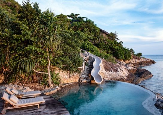 Pool mit Wasserrutsche, umgeben von Bäumen und Blick auf das Meer.