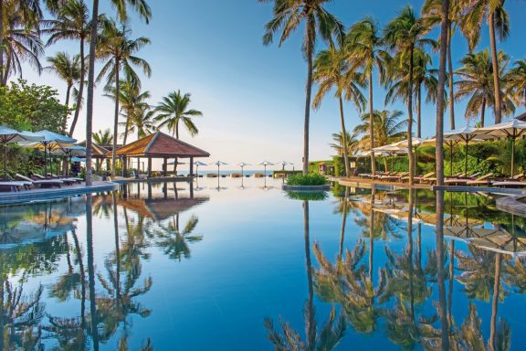 Tropischer Ort mit Palmen, Pool und Blick auf das Meer unter klarem Himmel.
