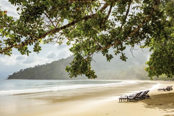 Strand mit Liegen unter einem Baum, sanfte Wellen und Berge im Hintergrund.