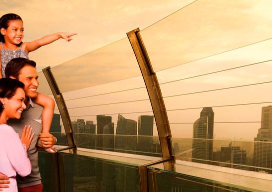 Familie steht an einem Balkon mit Aussicht auf eine Stadtlandschaft bei Sonnenuntergang.